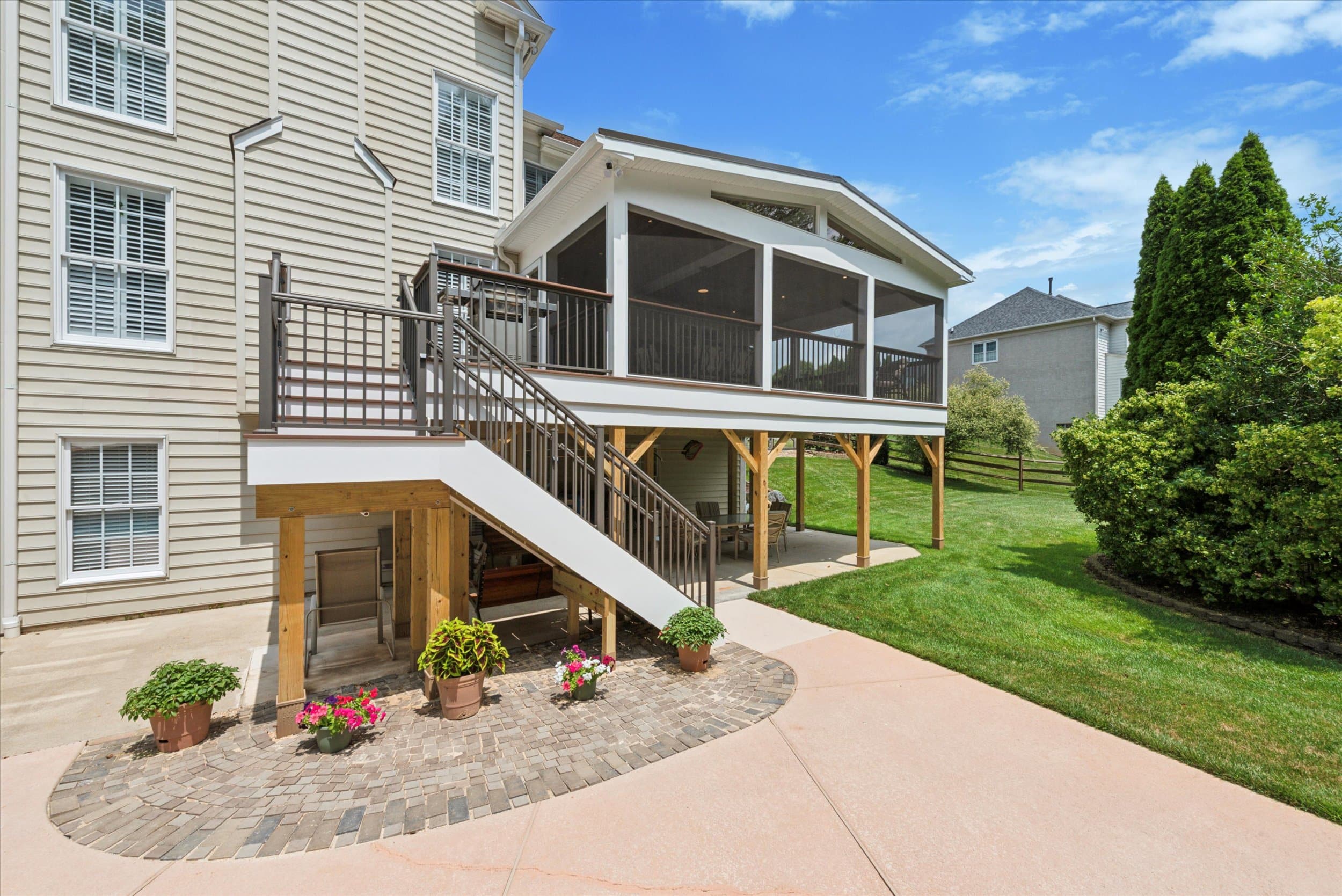 A beige house with white trim features a screened-in porch on a raised deck, accessible by stairs. Below is a paved patio with potted plants, surrounded by green grass and landscaping.