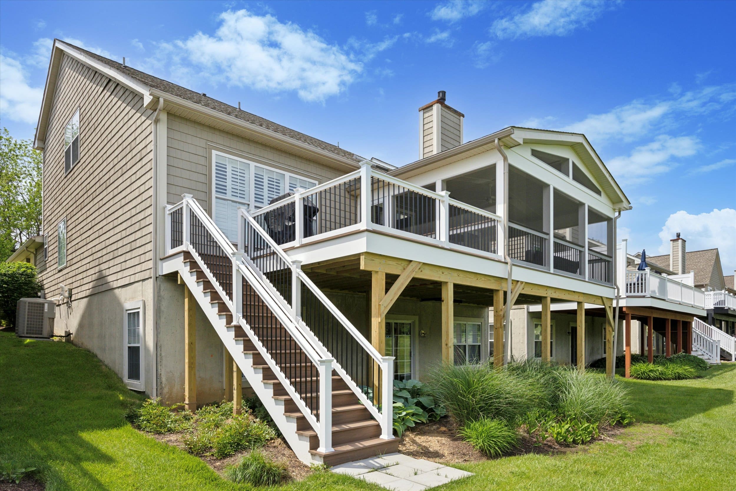 A tan house with a large elevated deck and screened porch, featuring white railings and stairs leading to a grassy backyard with landscaped plants, under a bright blue sky with scattered clouds.