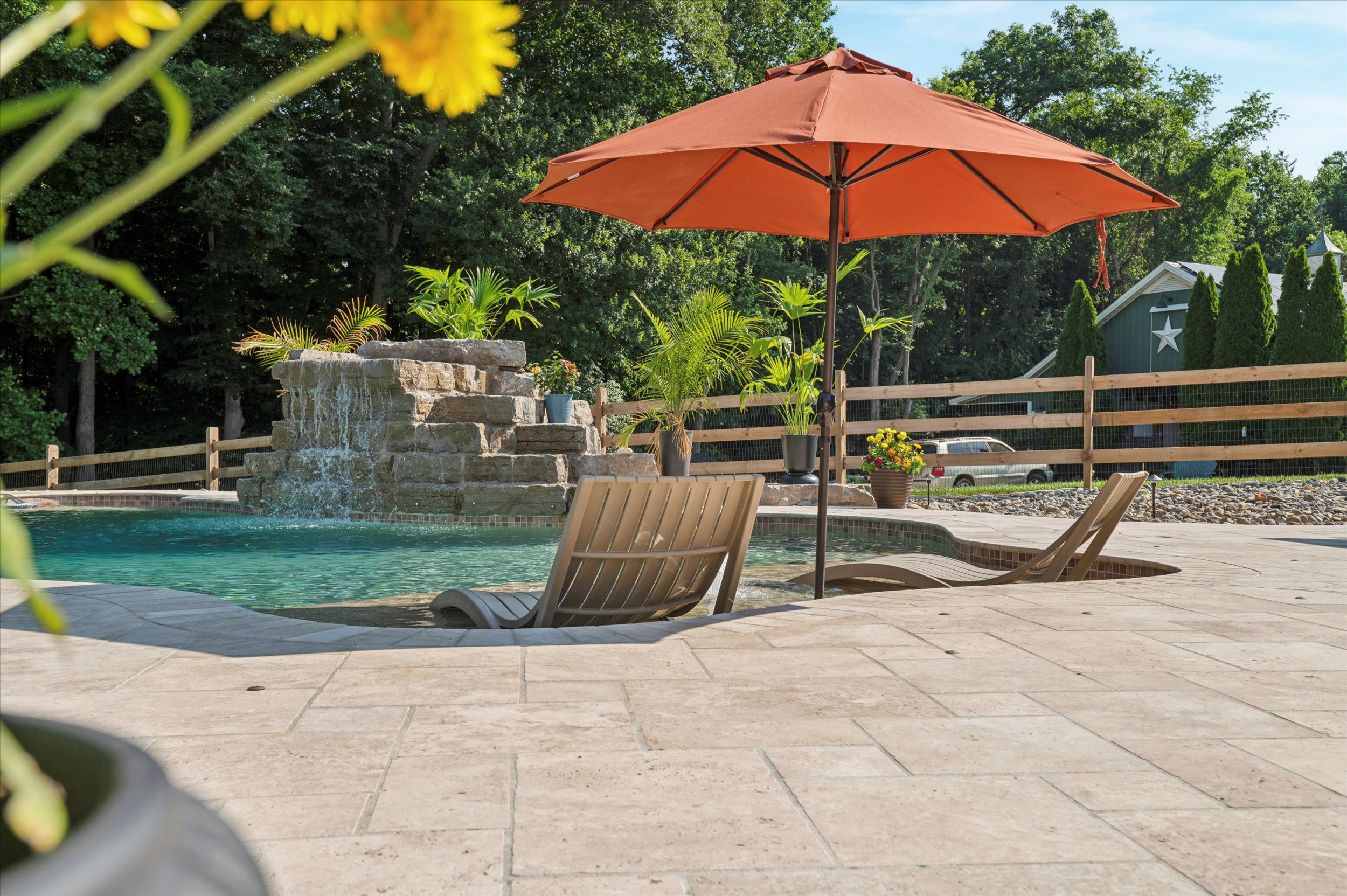 Two lounge chairs sit partially submerged in a pool beside an orange umbrella. A waterfall feature, potted plants, and a wooden fence are in the background, with trees and a house visible beyond the fence.