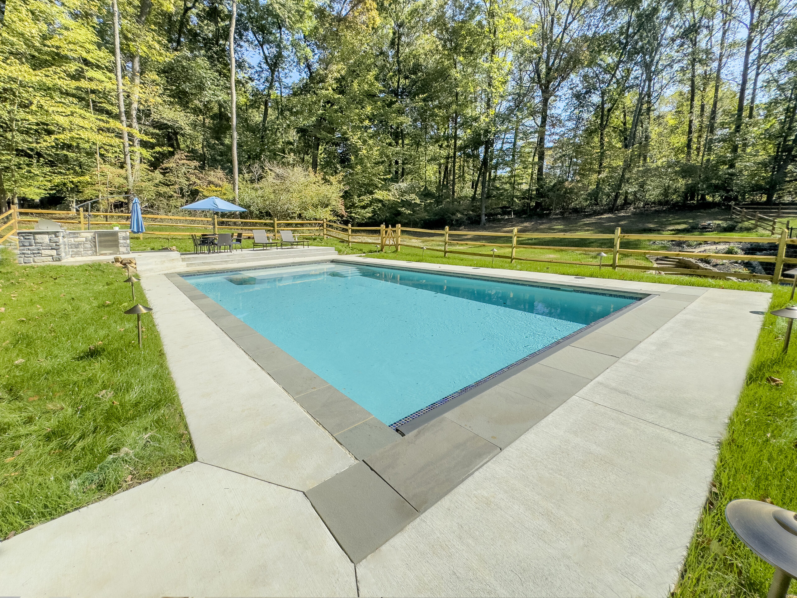 A rectangular outdoor swimming pool surrounded by a concrete deck, with green grass, wooden fence, patio table, and trees in the background on a sunny day.