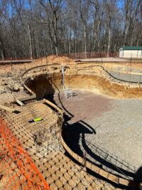 Image 2 | First State Building & Design A large, partially dug swimming pool construction site surrounded by orange safety fencing, with exposed dirt, gravel, and construction materials visible; a wooded area is in the background.