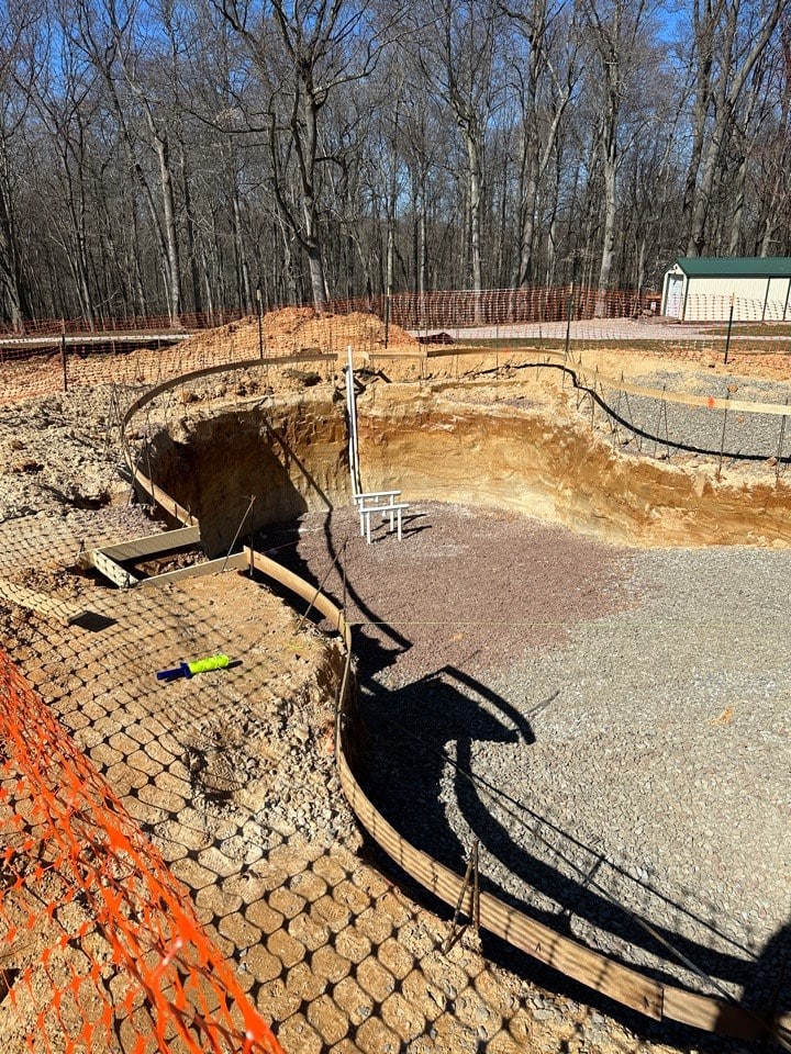 A large, partially dug swimming pool construction site surrounded by orange safety fencing, with exposed dirt, gravel, and construction materials visible; a wooded area is in the background.