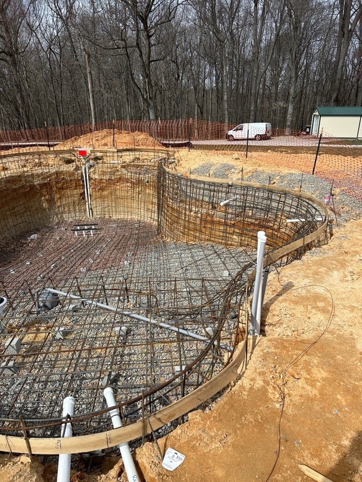 A swimming pool under construction with exposed rebar, pipes, and a dirt foundation, surrounded by bare trees, orange safety fencing, and a white van parked near a small building in the background.