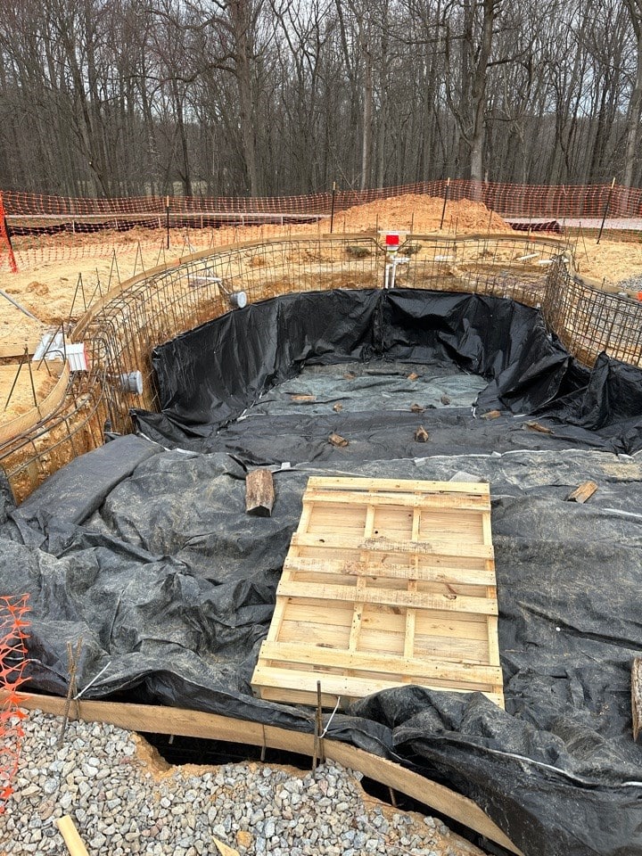 A construction site with a partially built in-ground pool lined with black plastic and rebar. A wooden pallet covers part of the pool. Orange safety fencing and bare trees are visible in the background.