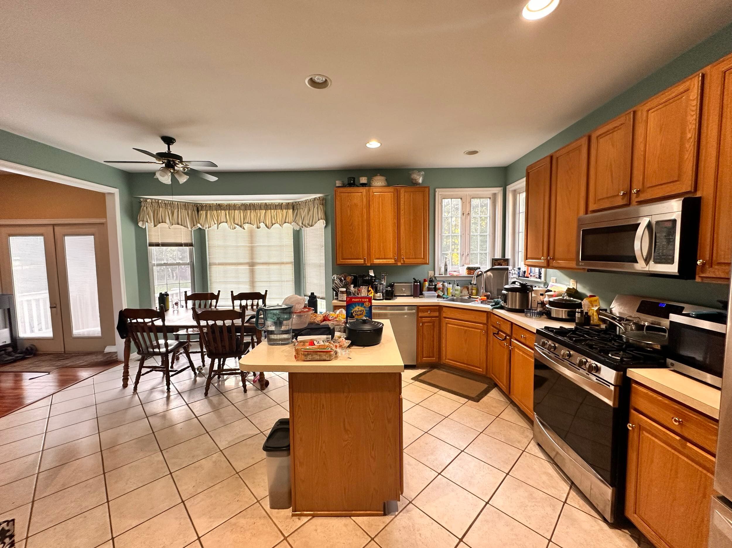 Photo-1 A spacious kitchen with wooden cabinets, stainless steel appliances, a central island, and a dining area with six chairs. The countertops are cluttered, and large windows let in natural light.