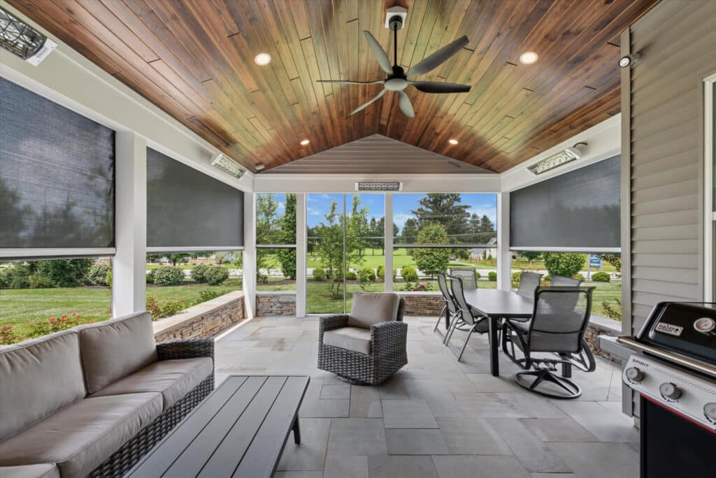 A modern screened-in porch with a wood-paneled ceiling, ceiling fan, outdoor sofa, coffee table, wicker chair, dining table with chairs, and a grill, overlooking a green, landscaped yard.