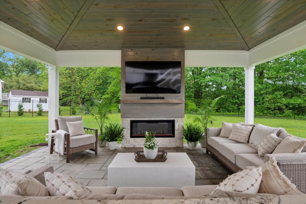 Covered outdoor patio with sofas, a white coffee table, potted plants, a modern fireplace, and a TV mounted above. The area features a wood-paneled ceiling and overlooks a green backyard with trees and grass.