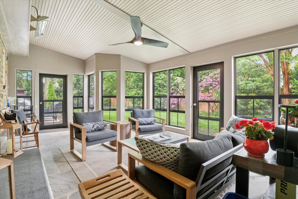 Bright sunroom with large windows, neutral walls, and a ceiling fan. Modern gray sofas with cushions, a coffee table, plants, and views of a green yard and blooming trees outside. Natural light fills the cozy space.