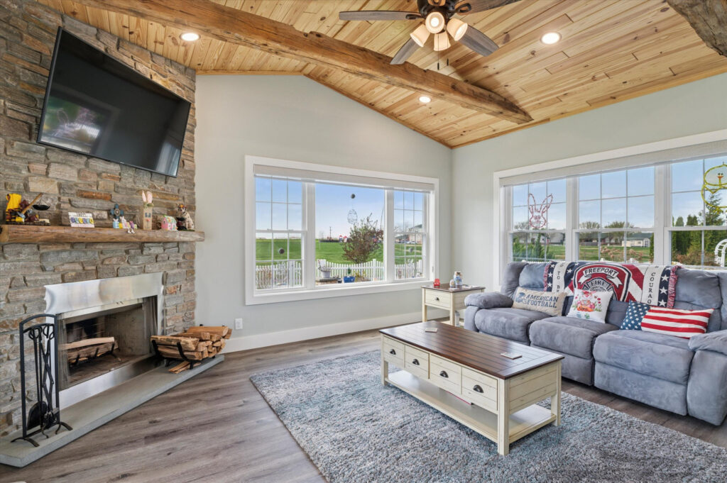 A cozy living room with a stone fireplace, mounted TV, gray sofa with pillows, wooden coffee table, and large windows offering a view of a green yard and partially cloudy sky. The ceiling is wood-paneled with exposed beams.