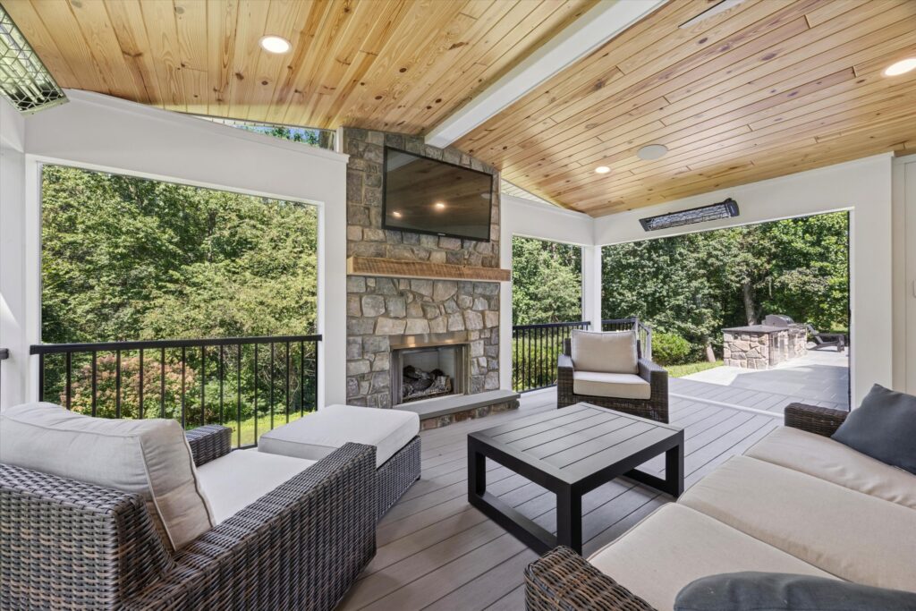 A covered outdoor patio with wicker furniture, a wood-paneled ceiling, ceiling lights, a stone fireplace with a mounted TV, and views of trees and a backyard grilling area.
