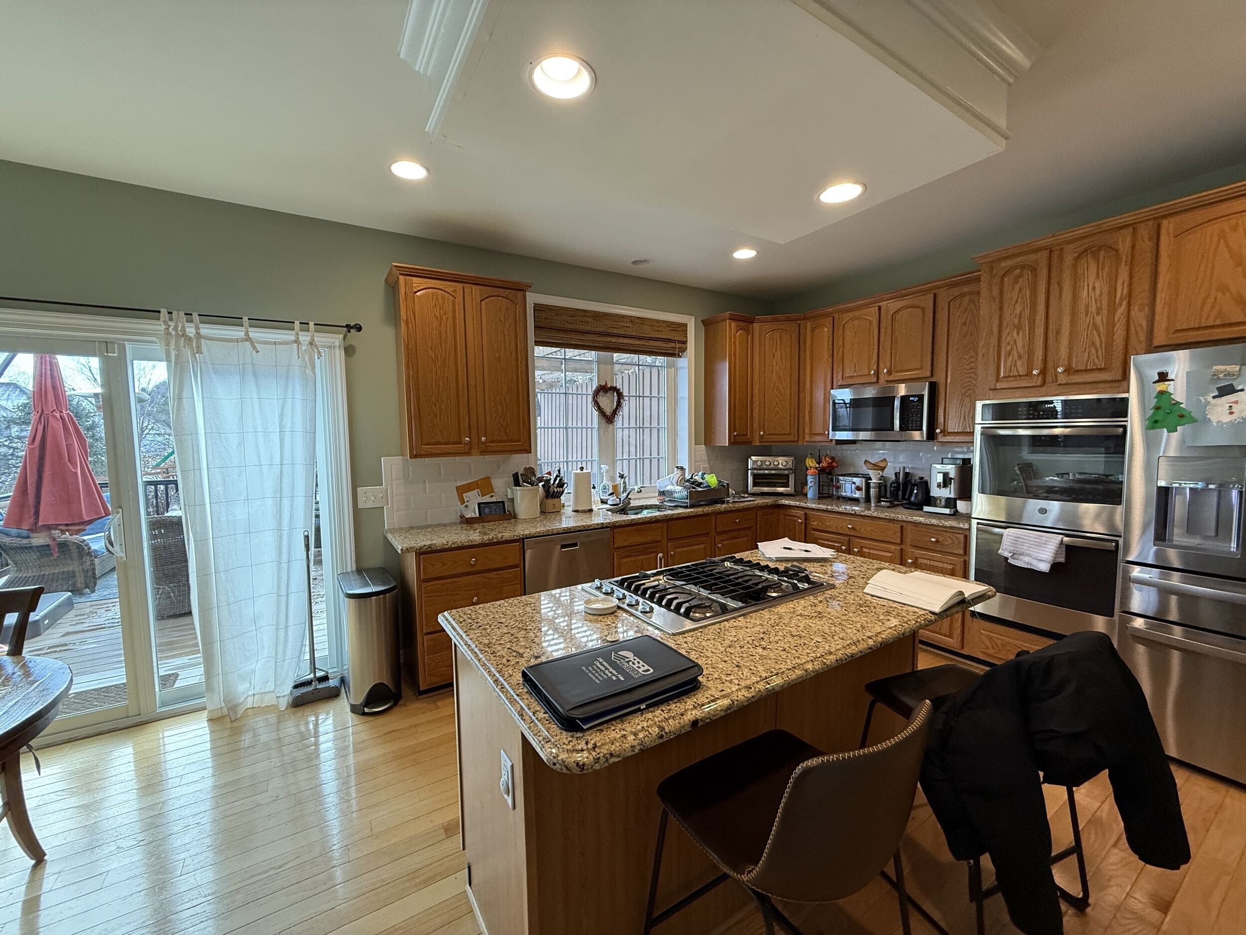 A spacious kitchen with wooden cabinets, stainless steel appliances, and a granite island countertop. Two chairs are at the island, and sliding glass doors lead to a patio with a red umbrella visible outside.