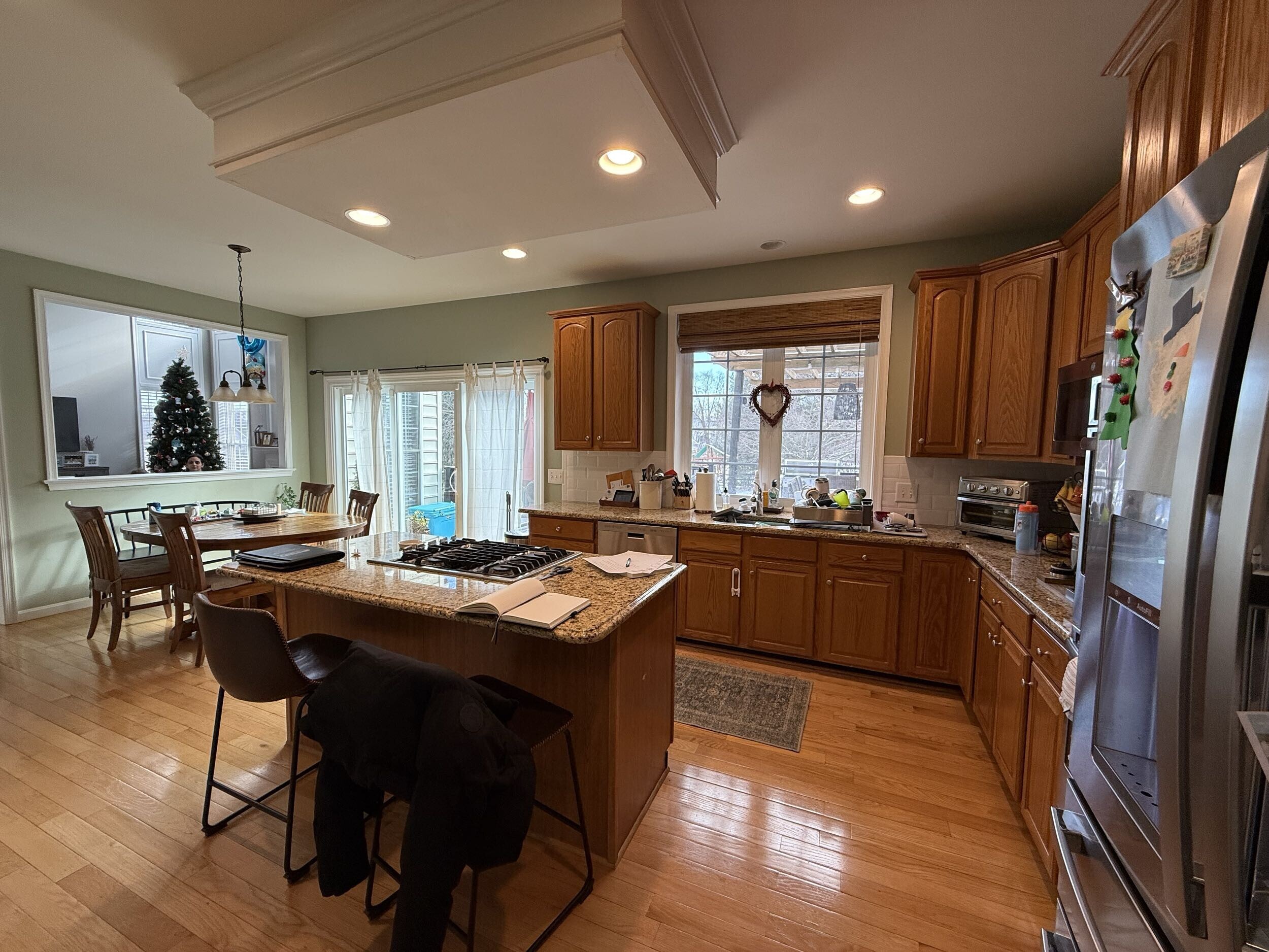 A spacious kitchen with wooden cabinets, an island with chairs, and stainless steel appliances. Sunlight enters through large windows and glass doors. A dining area with a Christmas tree is visible in the background.