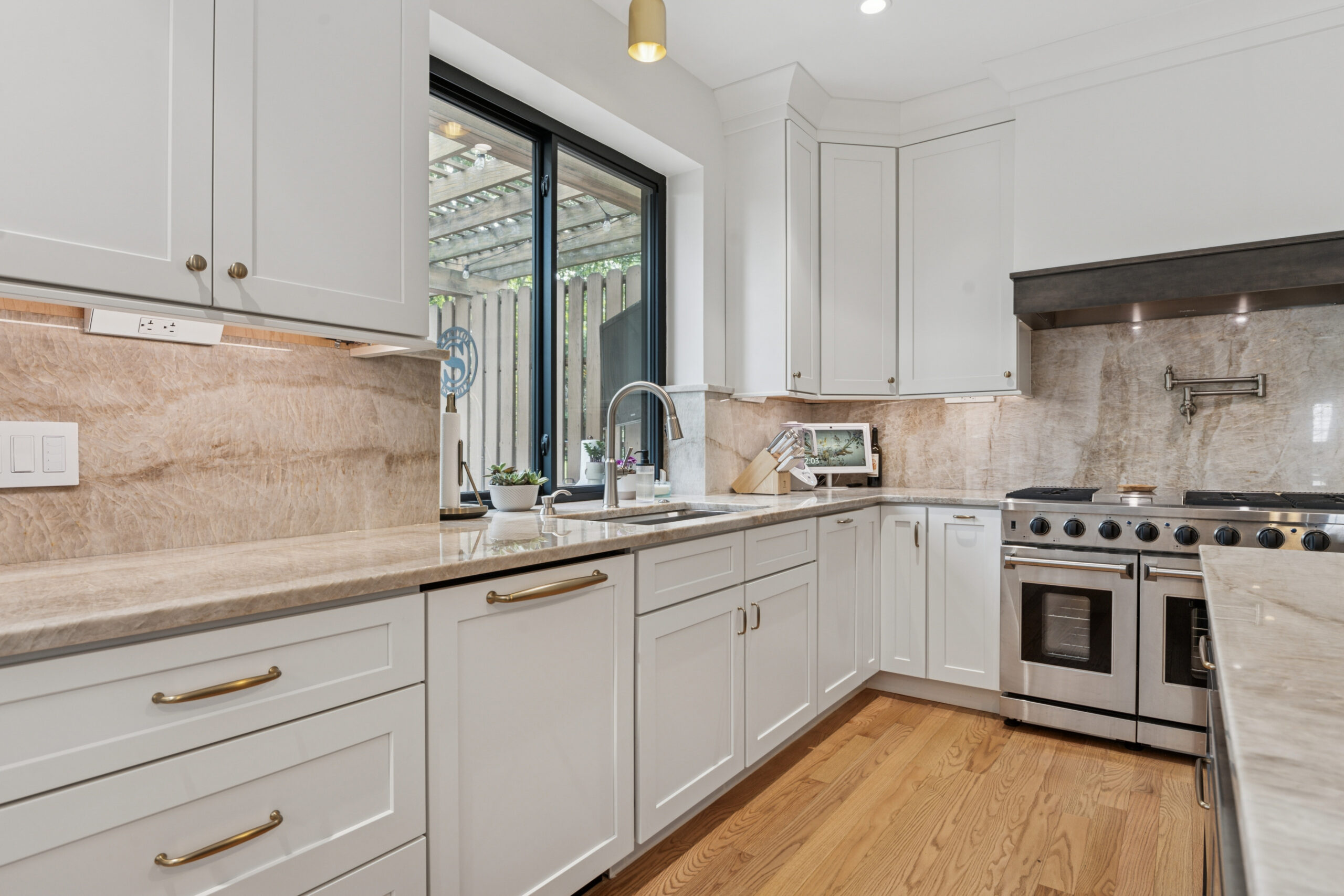 Final 1 Modern kitchen with white cabinets, stainless steel appliances, beige marble countertops and backsplash, wood flooring, and a large window overlooking a patio with a pergola.