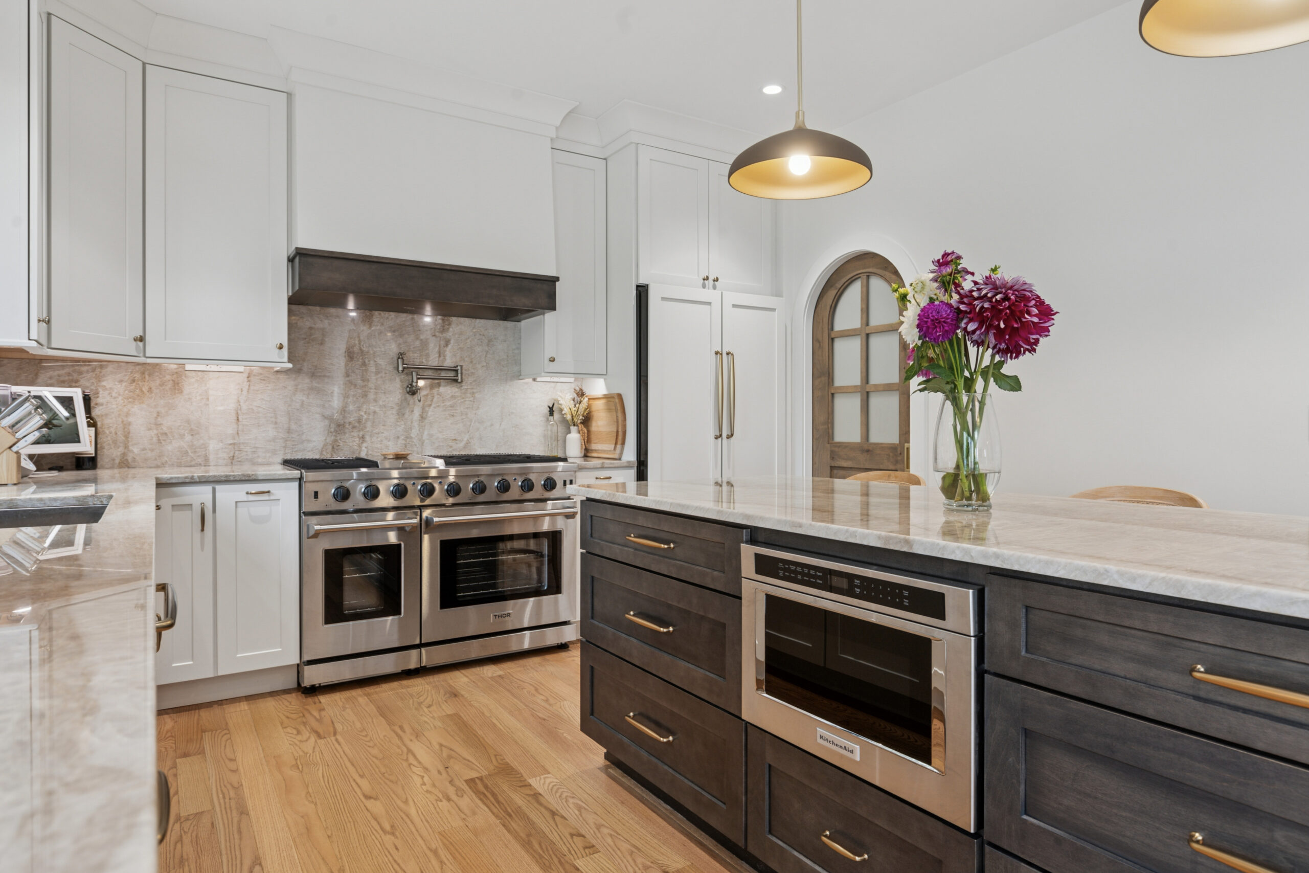 Modern kitchen with white cabinets, stainless steel appliances, dark wood island with built-in microwave, marble countertops, and a vase of purple flowers under pendant lights. Hardwood floor and arched doorway in the background.