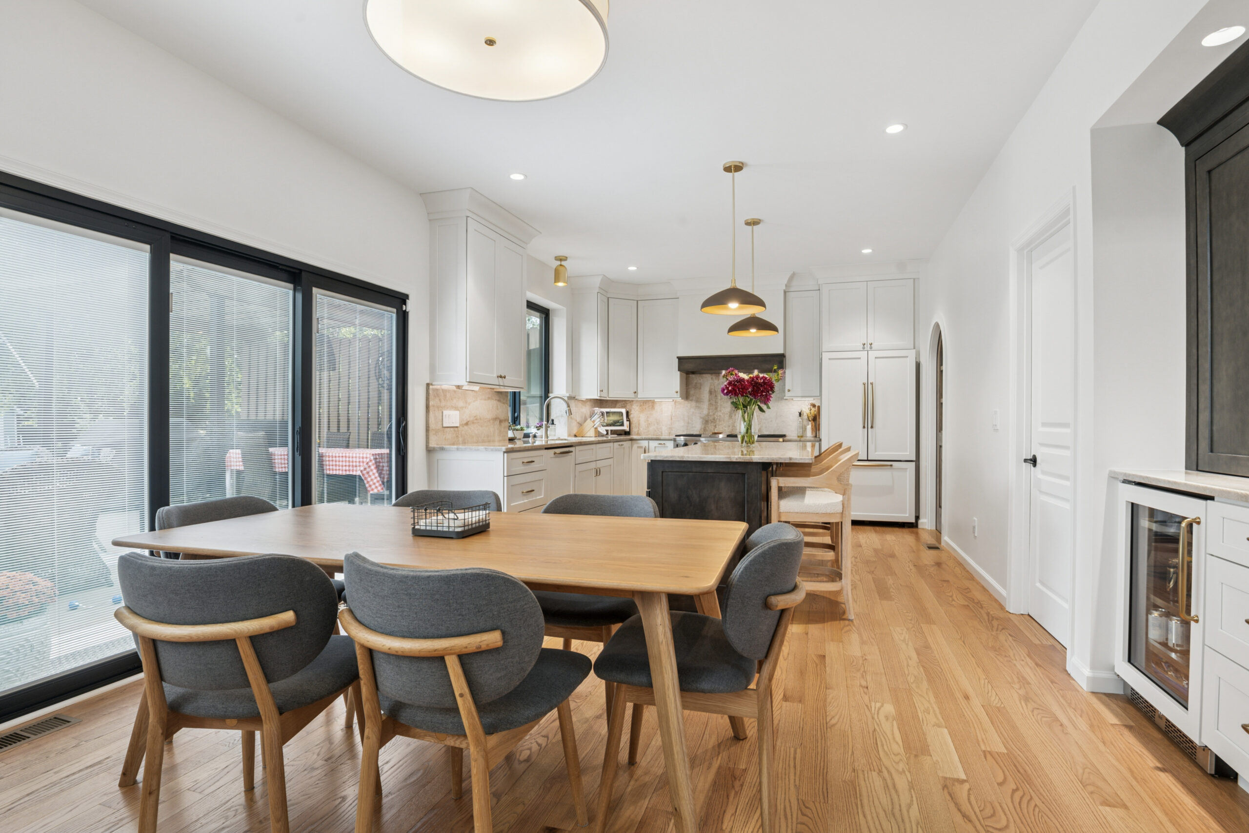 Modern kitchen and dining area with wooden floors, a wooden table with gray chairs, white cabinetry, a kitchen island with pendant lights, and large sliding glass doors leading outside.