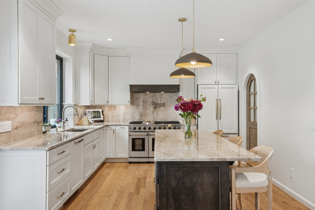 Modern kitchen with white cabinets, marble countertops, stainless steel appliances, a central island with flowers in a vase, wooden bar stools, pendant lights, and hardwood floors.