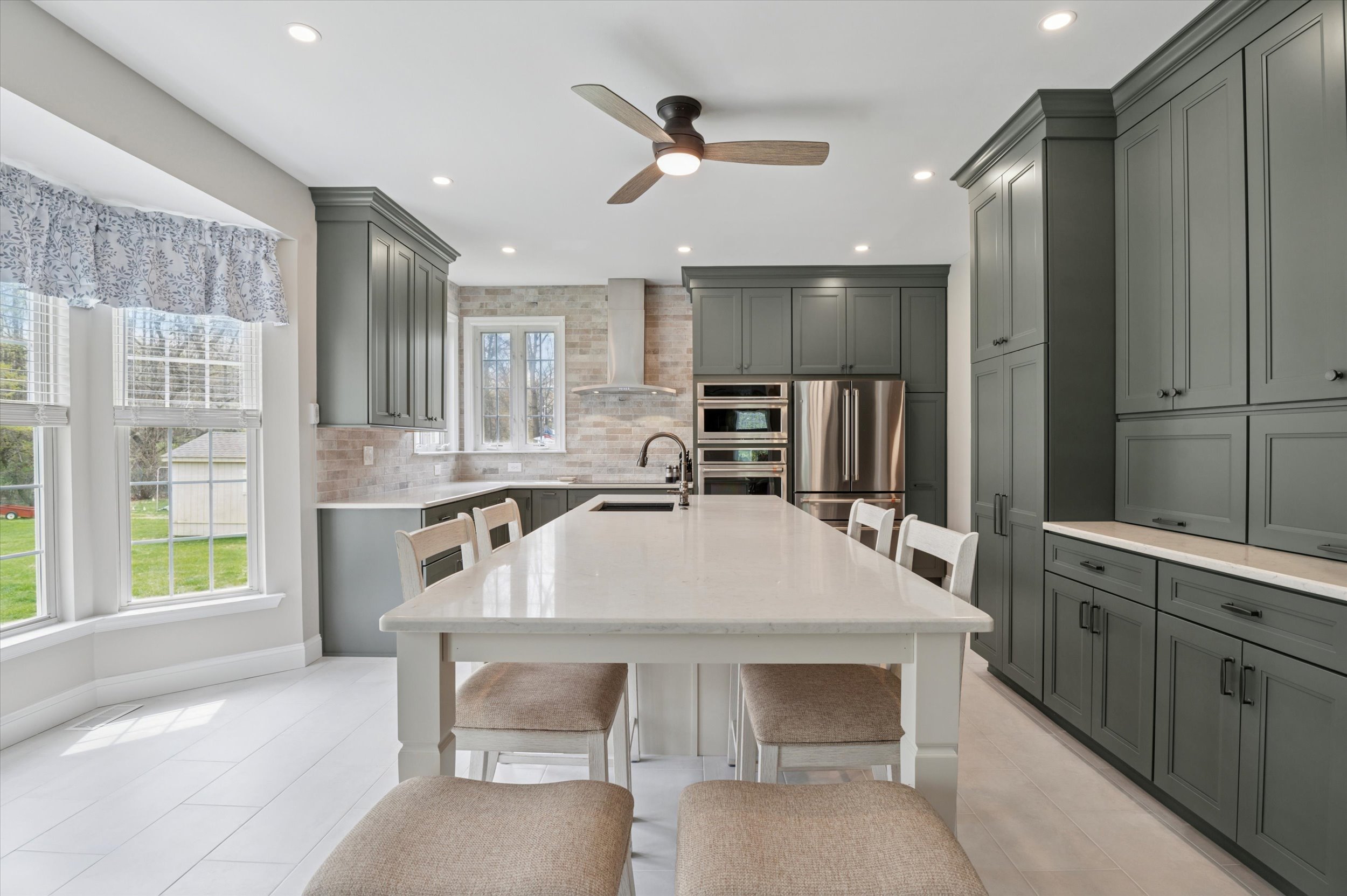 Interior-2 | First State Building & Design Modern kitchen with sage green cabinets, stainless steel appliances, a large white island with four beige chairs, tile backsplash, built-in oven, ceiling fan, and a window with a valance overlooking a green yard.