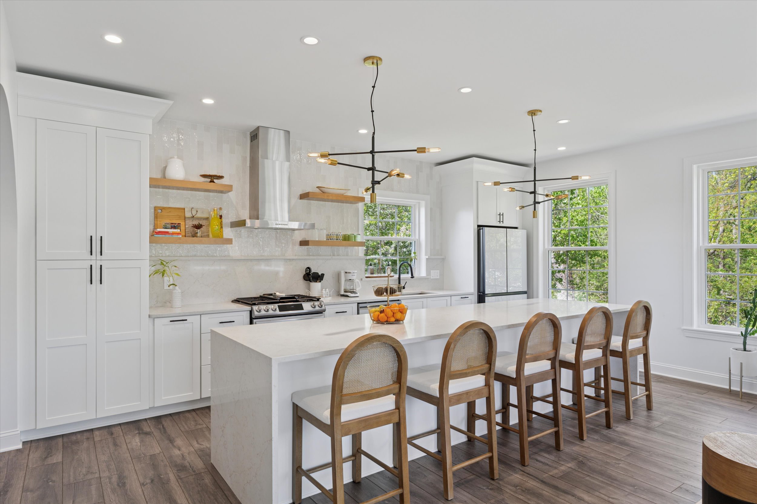 Modern kitchen with white cabinets, a large island with five wooden barstools, pendant lights, stainless steel appliances, open shelving, and large windows letting in natural light. A bowl of oranges sits on the island.