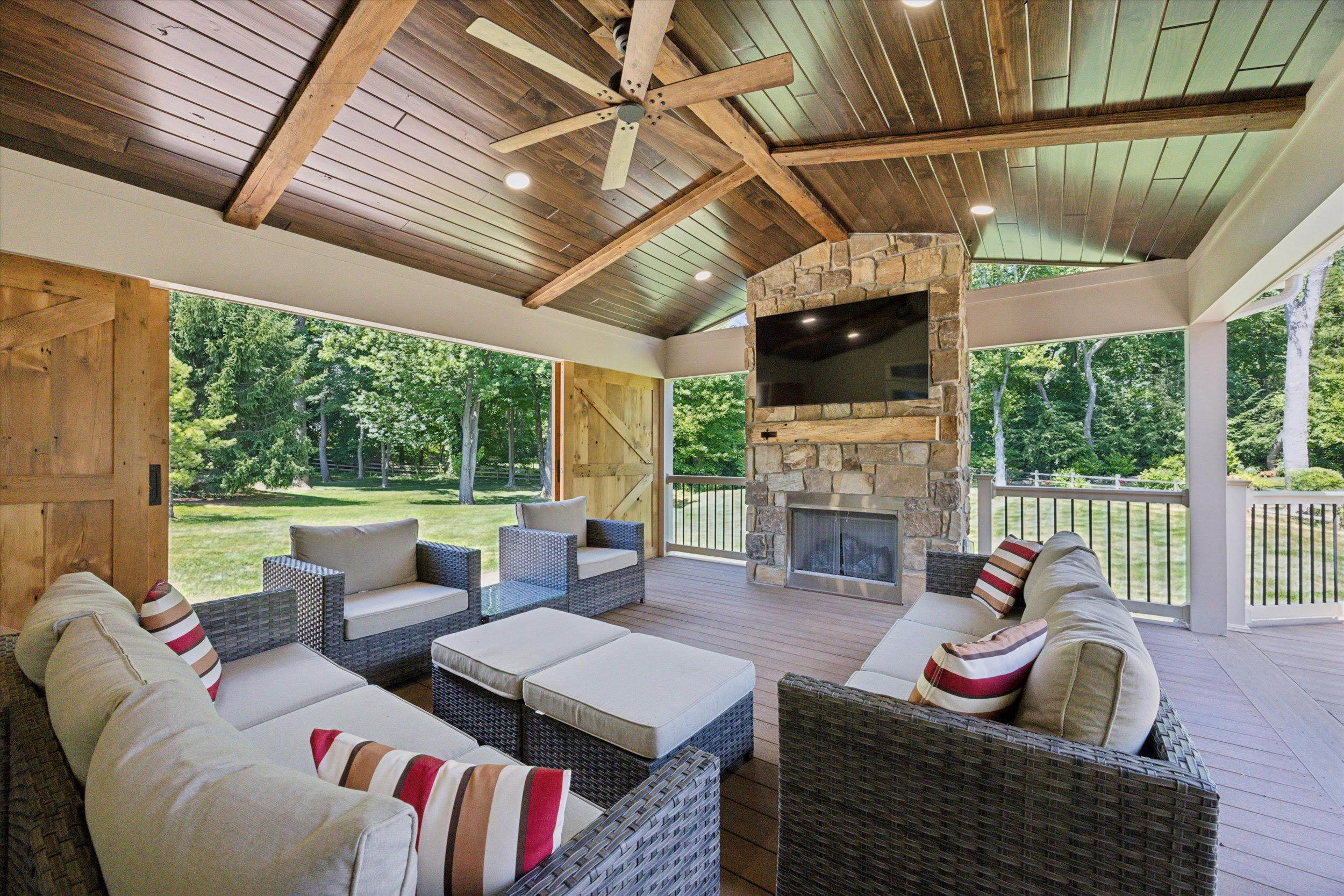 A covered patio with wicker sofas and chairs, striped cushions, a stone fireplace with a TV above it, wood ceiling beams, and a view of a green backyard through open sides.