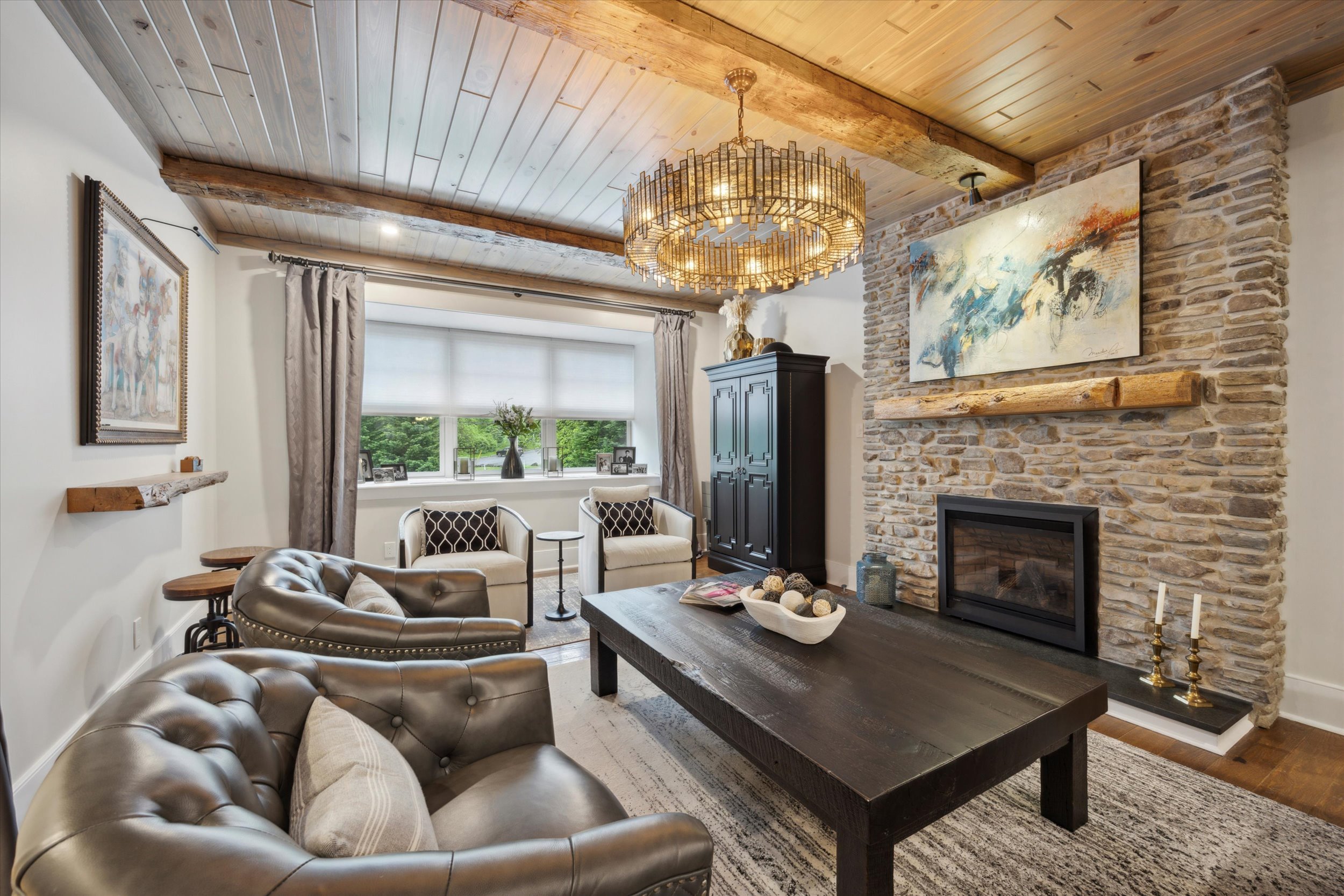 A cozy living room with tufted leather sofas, a dark wooden coffee table, a stone fireplace, and a large chandelier. There’s a window with gray curtains, artwork, and a television cabinet against the wall.