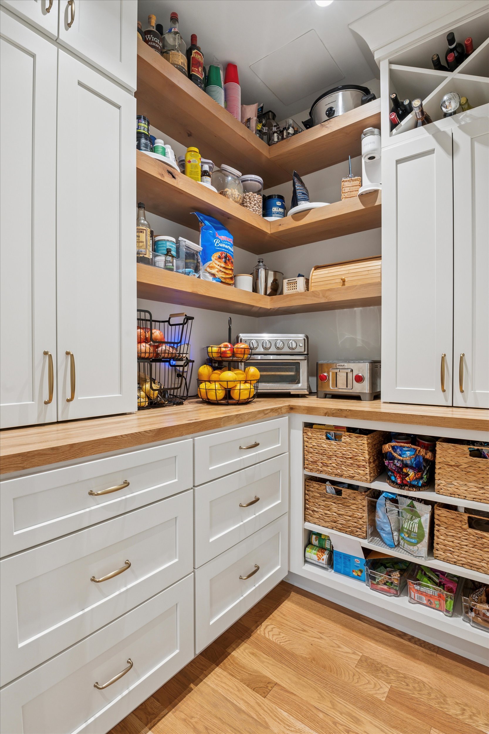 Beautifully Organized Walk-In Pantry With Custom Cabinetry and Wood Shelving A bright, organized walk-in pantry featuring white shaker cabinets, wooden countertops, open wood shelves with food and appliances, woven baskets, and neatly arranged produce on a hardwood floor.