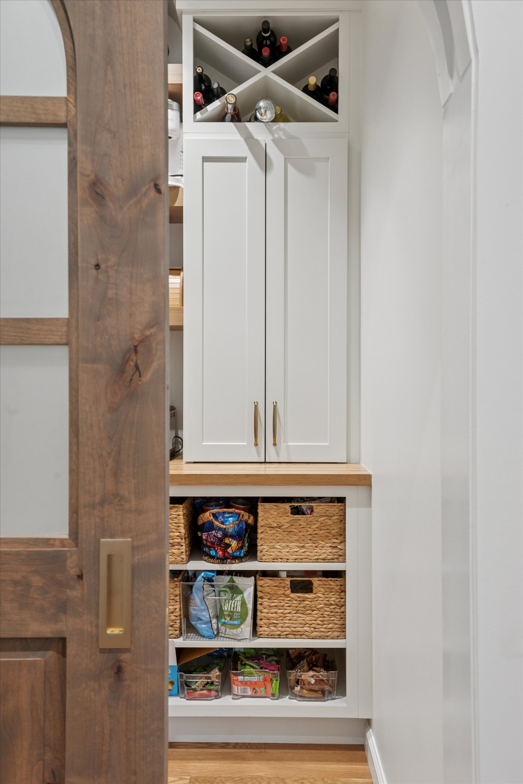 Custom Pantry Cabinetry with Wine Storage and Woven Basket Organization A built-in pantry cabinet with white shaker doors, a top wine storage cubby, wood countertops, and organized lower shelves with woven baskets and clear storage bins.