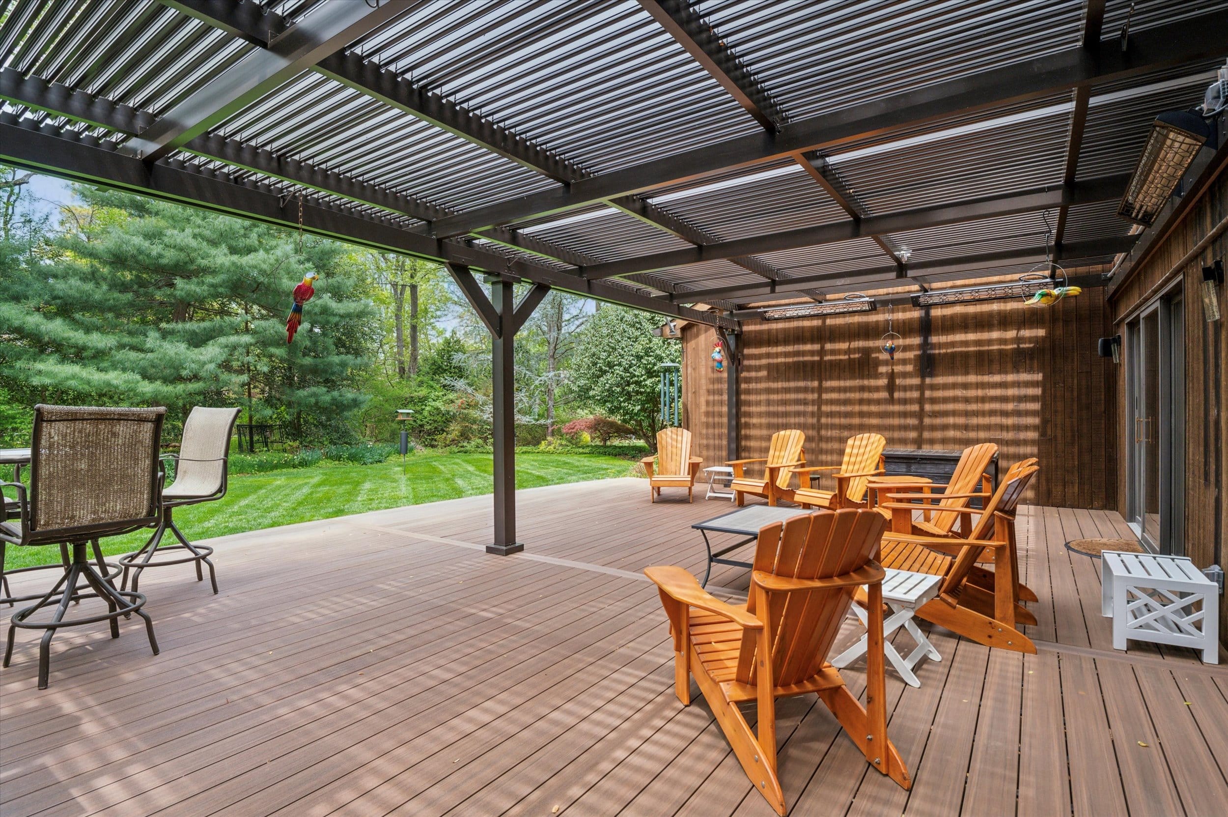 01-100 Santomera Ln_01 Outdoor wooden deck under a metal pergola with orange Adirondack chairs facing a green backyard garden.
