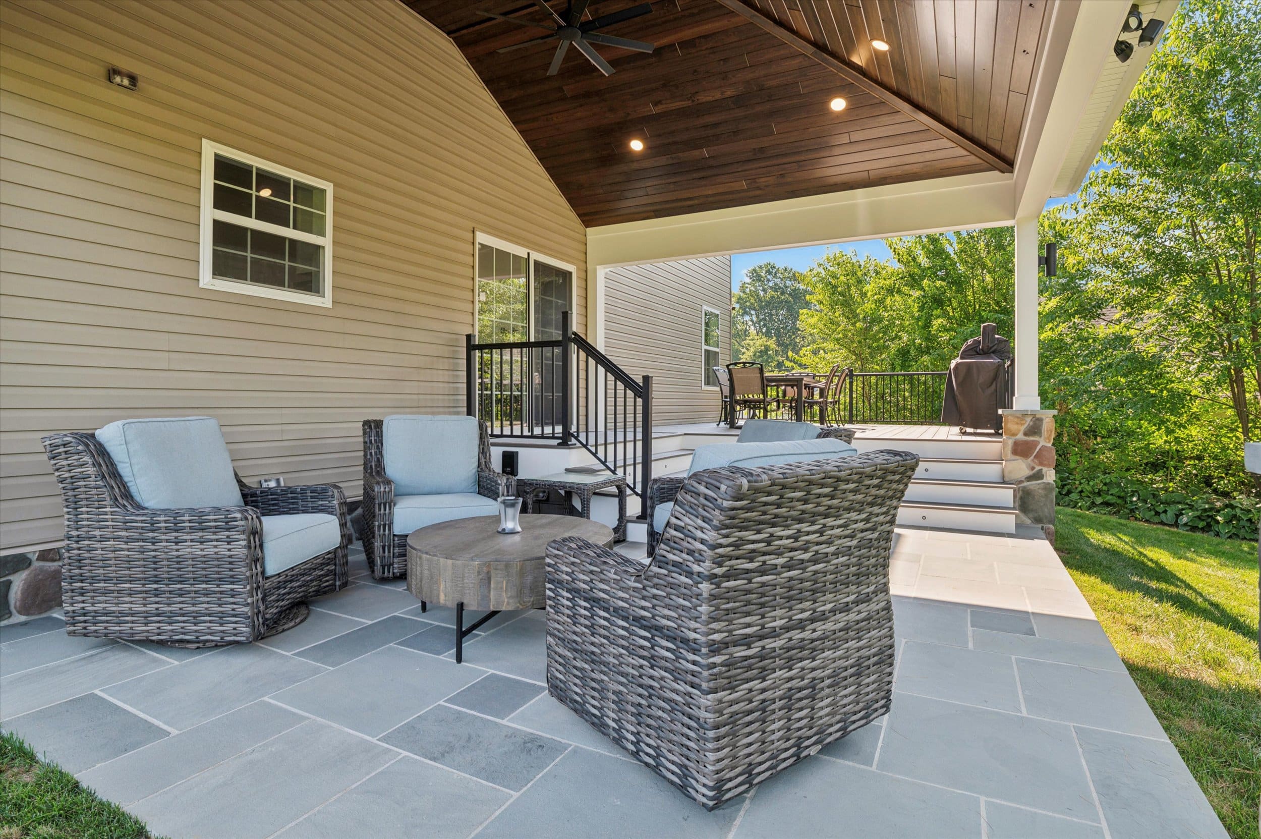 COD1E9_1 1 Cozy outdoor patio with woven grey-cushioned chairs around a round wooden coffee table on a tiled deck under a wooden ceiling fan. A dining table and grill are visible further back.