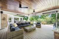 decks4 | First State Building & Design Covered outdoor deck with gray wicker sectional, yellow cushions, and a coffee table, overlooking a lush tree-filled view through a glass railing.