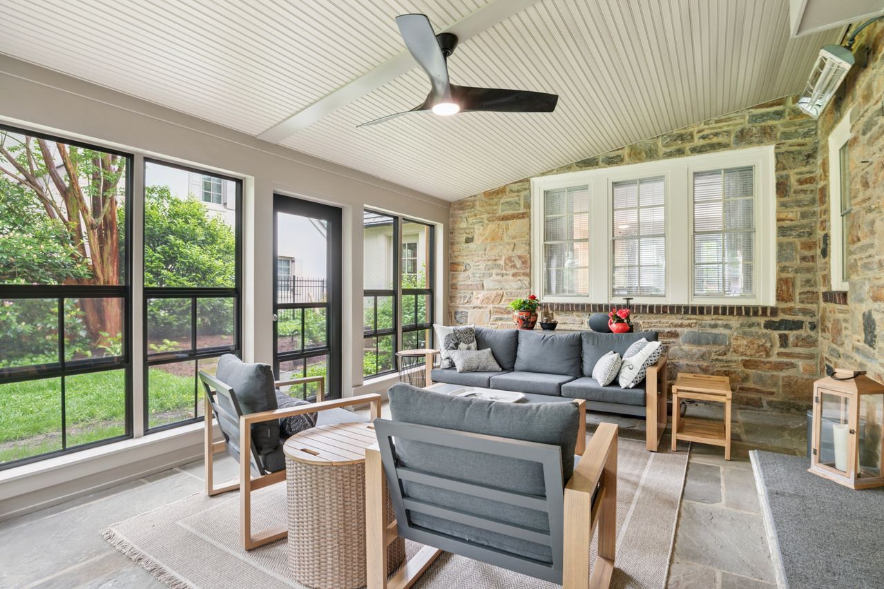 Cozy sunroom with gray cushioned seating, stone wall, and large windows showing greenery on a sunny day