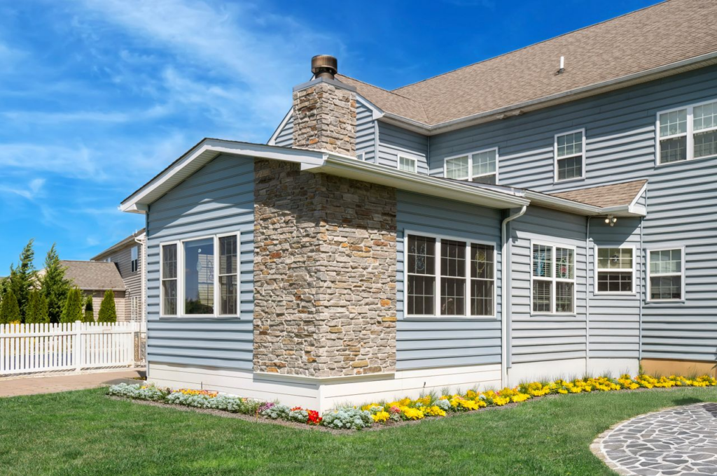 Blue suburban house with stone chimney, white-framed windows, and flower bed along the foundation.